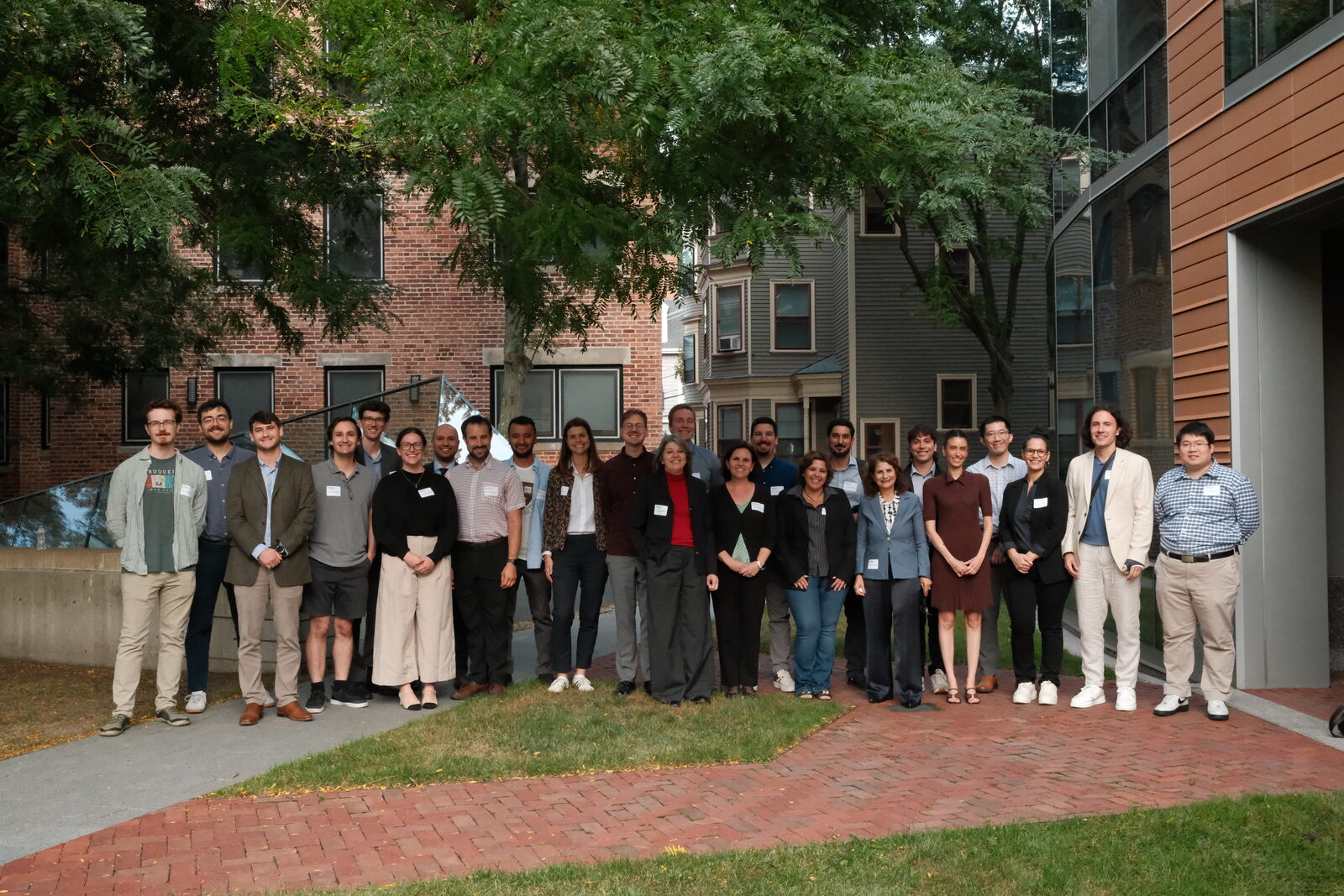 Group photo of about 25 conference participants standing outdoors on a brick path in front of trees and campus buildings.